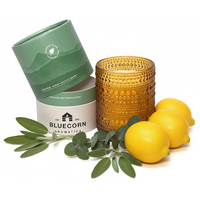 An amber glass candle with a textured dimple finish sits next to its light green and white cylindrical packaging surrounded by fresh greenery and lemons with a white background