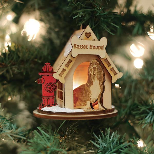 A wooden Christmas tree ornament shaped like a doghouse with Basset Hound on top, a carved image of a basset hound inside, and a red fire hydrant decoration beside it, hanging on a tree branch.