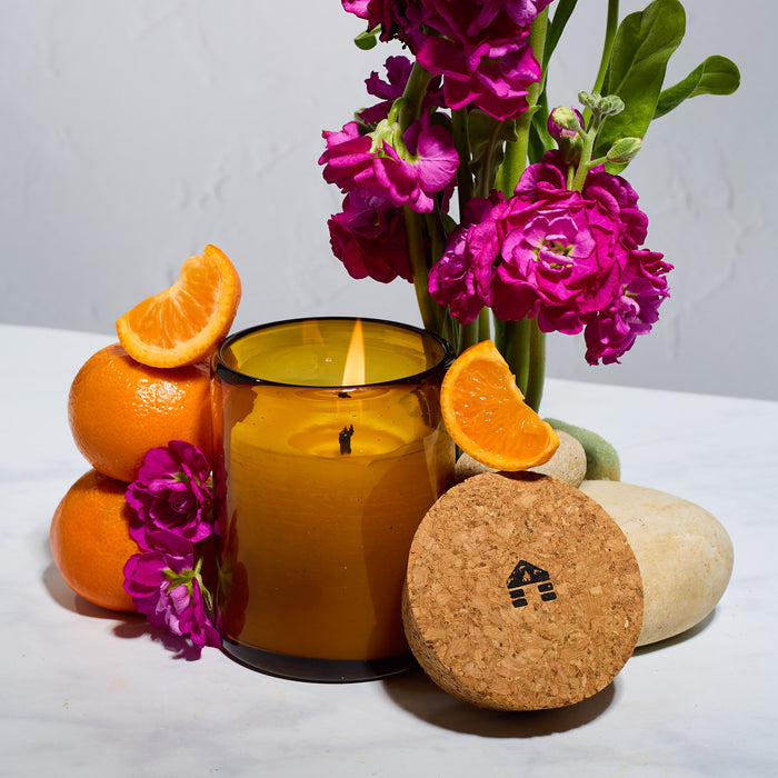 A lit bluecorn beeswax candle in a brown glass jar with orange slices, pink flowers and rocks on a white surface