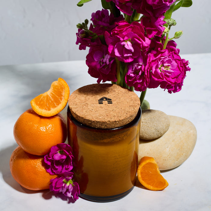 A  bluecorn beeswax candle in a brown glass jar with cork lid surrounded by orange slices, pink flowers and rocks on a white surface