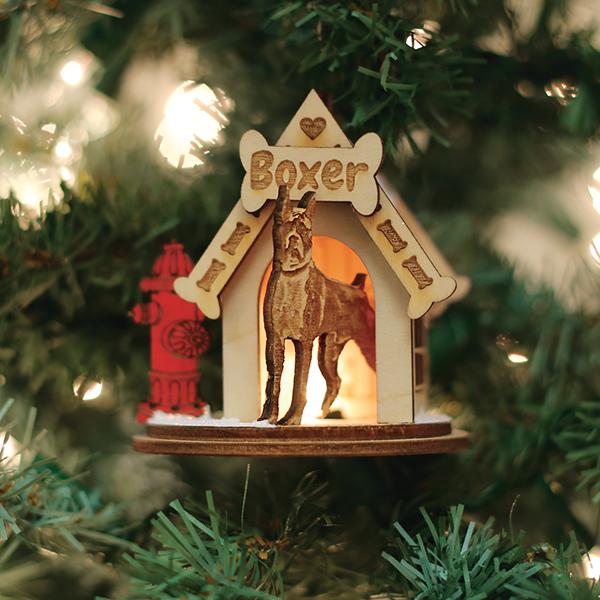 A wooden Christmas tree ornament shaped like a doghouse with a Boxer sign and a carved Boxer dog inside, hanging on a tree with lights. A small red fire hydrant decoration is attached to the side.