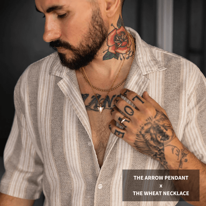 Man wearing a striped linen shirt with a layered gold arrow pendant and wheat necklace from the Build Your Own Bundle - Gold collection, showcasing bold men's jewelry with detailed tattoos visible on neck and hand, styled in a confident, luxurious fashion.