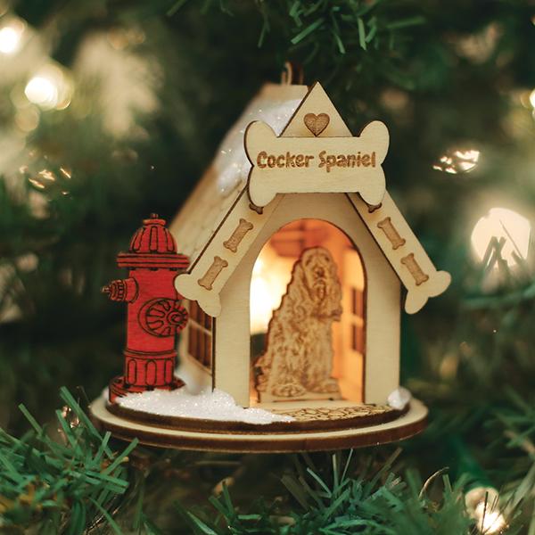 A wooden Christmas tree ornament shaped like a doghouse with a bone-shaped sign reading Cocker Spaniel, a red fire hydrant on the side, and a carved Cocker Spaniel dog inside, surrounded by green tree branches.