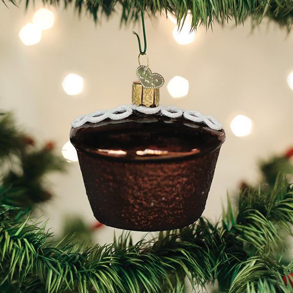 A Christmas tree ornament shaped like a cupcake with a gold top and white icing sits among green pine branches, with blurred lights in the background.