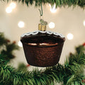 A Christmas tree ornament shaped like a cupcake with a gold top and white icing sits among green pine branches, with blurred lights in the background.