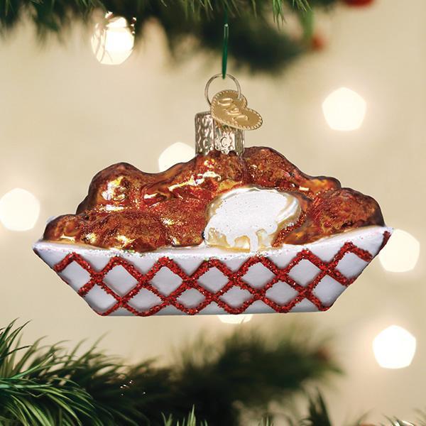 A Christmas tree ornament shaped like a tray of fried chicken with a dollop of mashed potatoes and gravy, decorated with red lattice detail, hangs on a tree with blurred lights in the background.