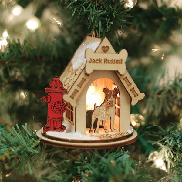 A Christmas tree ornament shaped like a doghouse with a Jack Russell figure inside, a red fire hydrant outside, and warm light glowing from within, hanging on a decorated tree.