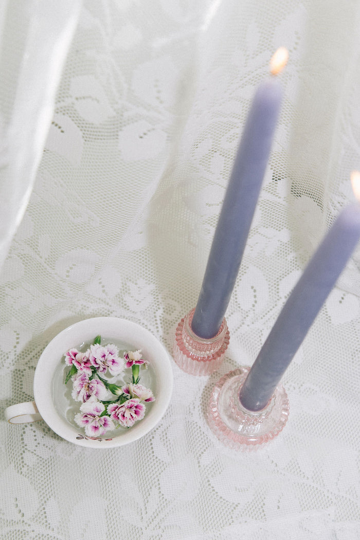 Two lit purple beeswax taper candles in decorative holders on a lace tablecloth with a small white dish featuring pink flowers.