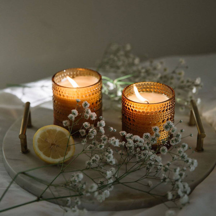 Two lit candles in textured glass holders on a marble tray with flowers and a lemon.