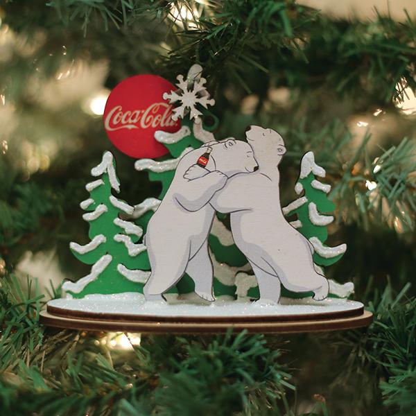 A Christmas tree ornament features two white polar bears hugging, with snow-covered pine trees and a red Coca-Cola ball in the background, hanging among green tree branches.