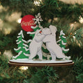 A Christmas tree ornament features two white polar bears hugging, with snow-covered pine trees and a red Coca-Cola ball in the background, hanging among green tree branches.