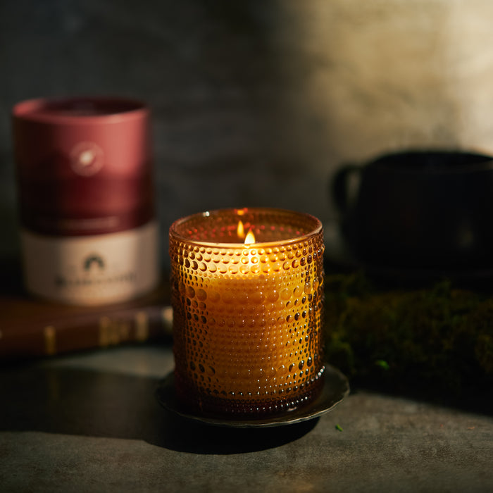 Fall scented candles glowing in a textured glass holder with a dark background and surrounding objects.