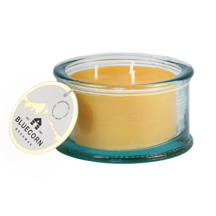 Image displays Bluecorn Beeswax 3-Wick pure beeswax candle in 100% recycled Spanish glass. The wax is golden in color and the glass is a watery aqua color. Candle has hang tag with Bluecorn name and logo and an outline of the mountains in gold foil.