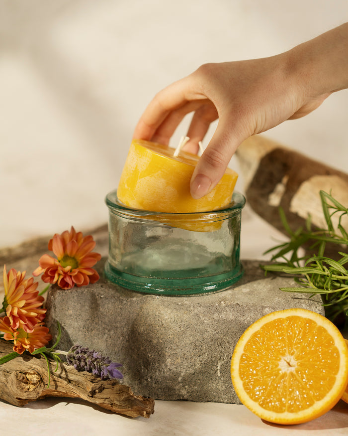 A 100% recycled 3-wick Spanish glass candle jar on top of a stone slab with a pure beeswax refill being inserted into the glass surrounded by flowers and orange slices
