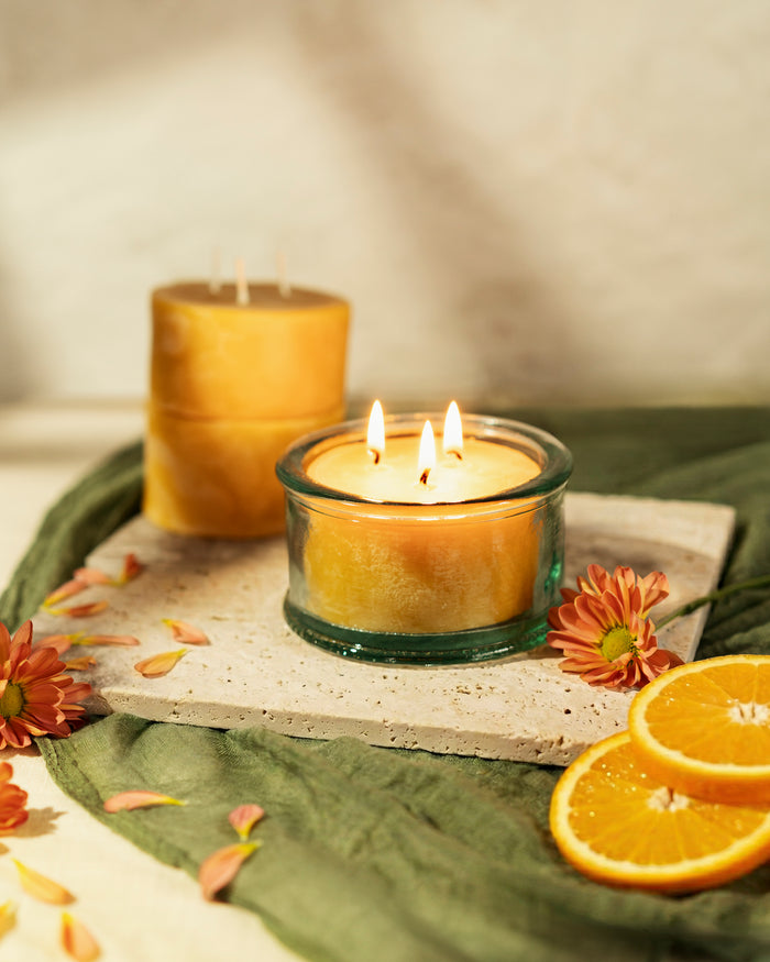 A 100% recycled 3-wick Spanish glass filled candle jar burning on top of a stone slab. There are two refill candles sitting next to the burning candle that are surrounded by orange flowers and orange slices with a neutral background