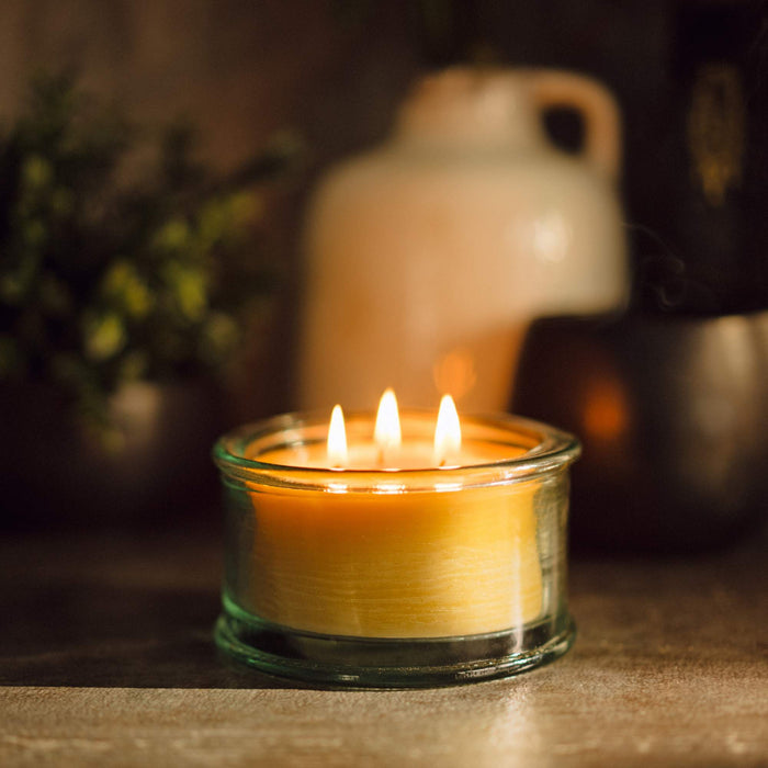 A 100% recycled 3-wick Spanish glass filled candle jar burning on top of a table with a milk jug and greenery in the background