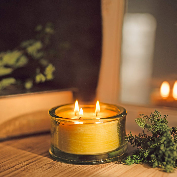 A 100% recycled 3-wick Spanish glass filled candle jar burning on top of a wooden table with a closed book and greenery in the background