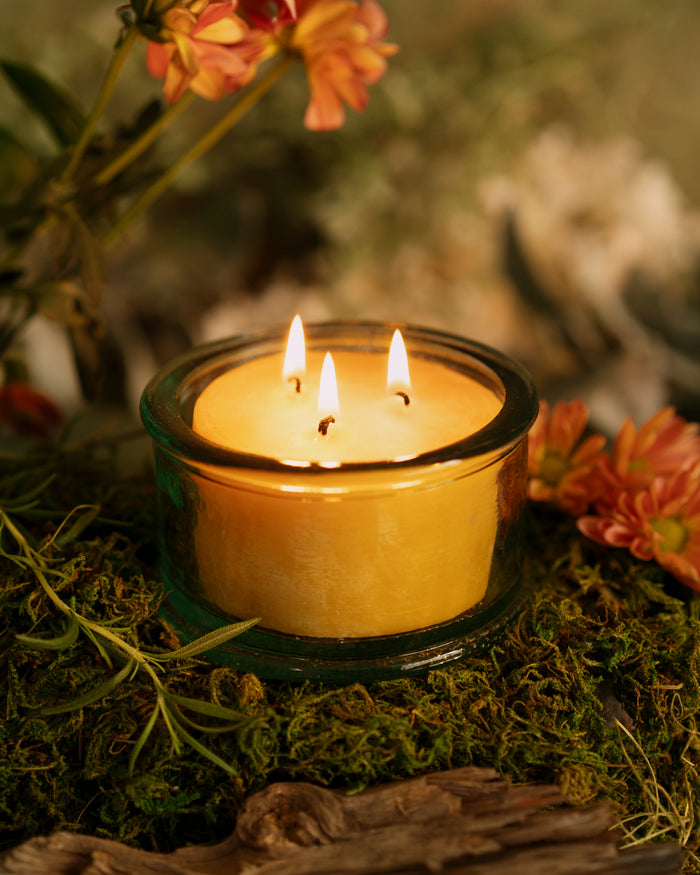 Beeswax scented candle in handmade recycled glass, featuring three wicks and surrounded by flowers and greenery.