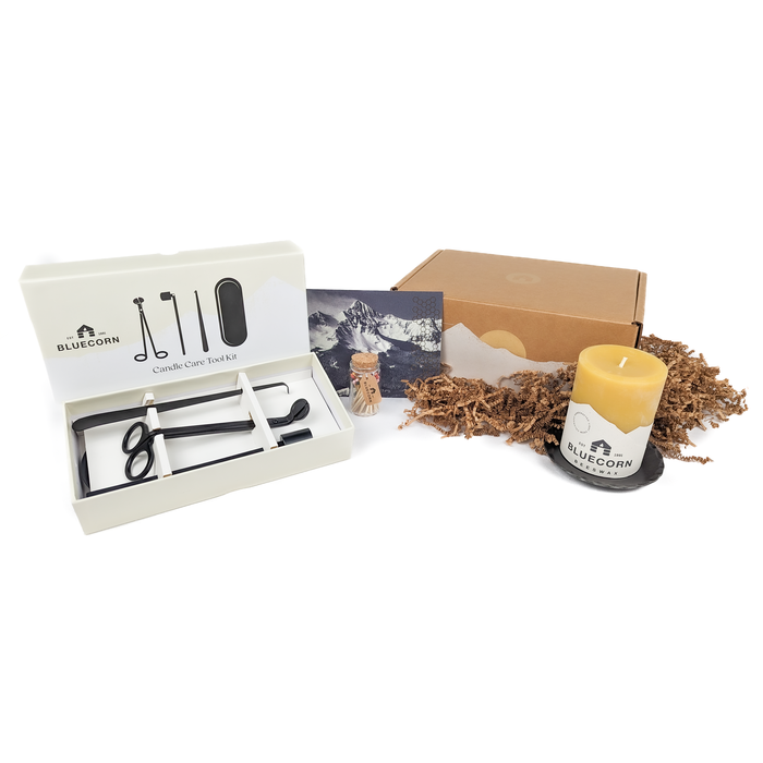 A Pure Beeswax Pillar Candle Gift Set with a gold-foiled gift box, including a 3" x 4" pure beeswax pillar candle, a 4.75" hand-forged pillar base, a candle care toolkit, a refillable apothecary jar of rainbow matches, and a gift card with a handwritten message.
