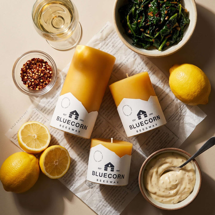 3 Bluecorn Beeswax raw pillar candles with white bluecorn label with lemons, a bowl of greens and spices and a glass of white wine