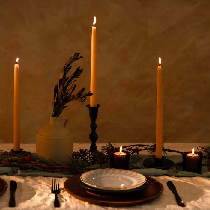 Three lit raw beeswax taper candles in taper candle holders in dinner table setting with holiday decor on a textured wall background