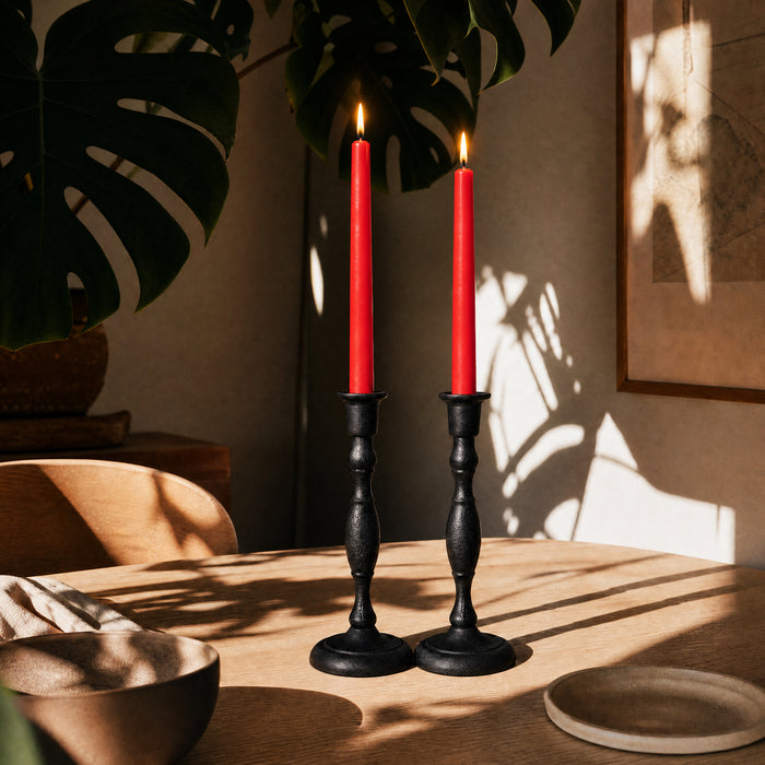 Two red beeswax taper candles in black candlesticks on a wooden table with a plant and wall art in the background.