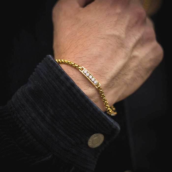 The Atlas Ice Bracelet on a man's wrist featuring a gold-toned chain with a rectangular bar set with clear cubic zirconia stones, styled with a dark textured jacket sleeve, bold by design.