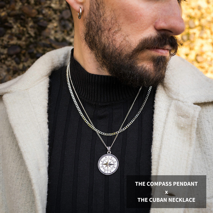 The Compass Essentials Bundle - Silver features a bold men's jewelry set with a silver compass pendant on a sleek Cuban chain necklace, styled with a black ribbed turtleneck and beige coat.