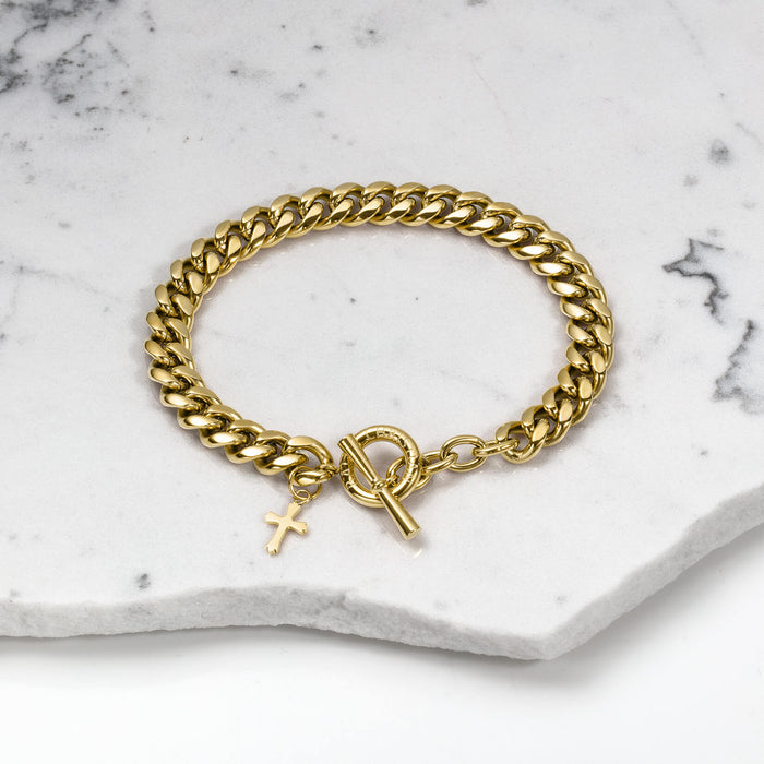 The Cross Pendant Bracelet - Gold features a polished gold chain with a small cross charm and a toggle clasp, displayed on a white marble surface, bold by design.
