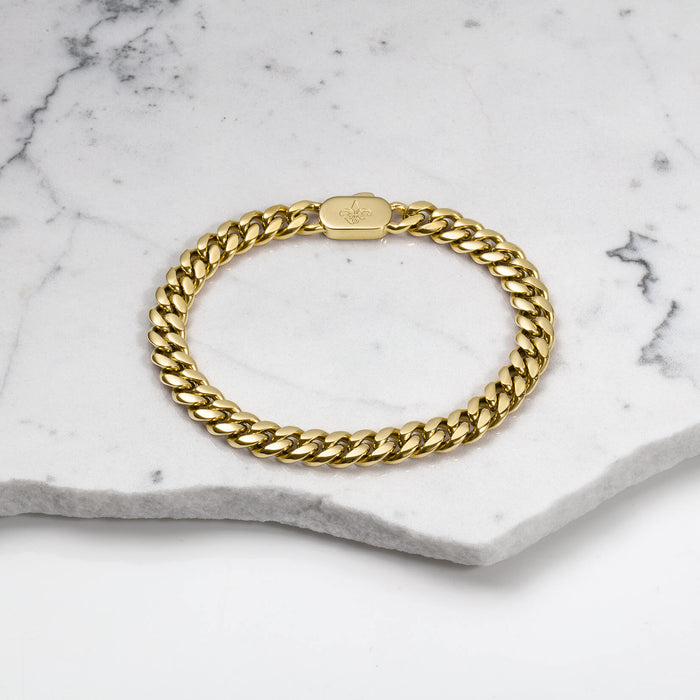 The Cuban Bracelet - Gold displayed on a white marble surface, featuring a polished, thick gold chain link design with a secure clasp engraved with a subtle logo.