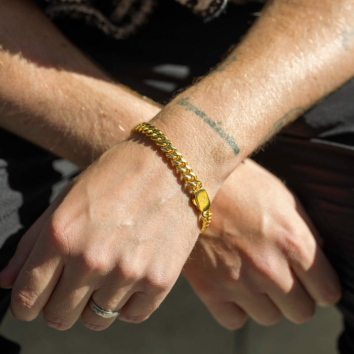 Close-up lifestyle shot of a wrist wearing The Cuban Bracelet - Gold, a bold and polished gold chain bracelet designed to be noticed, paired with casual attire and a tattooed arm.
