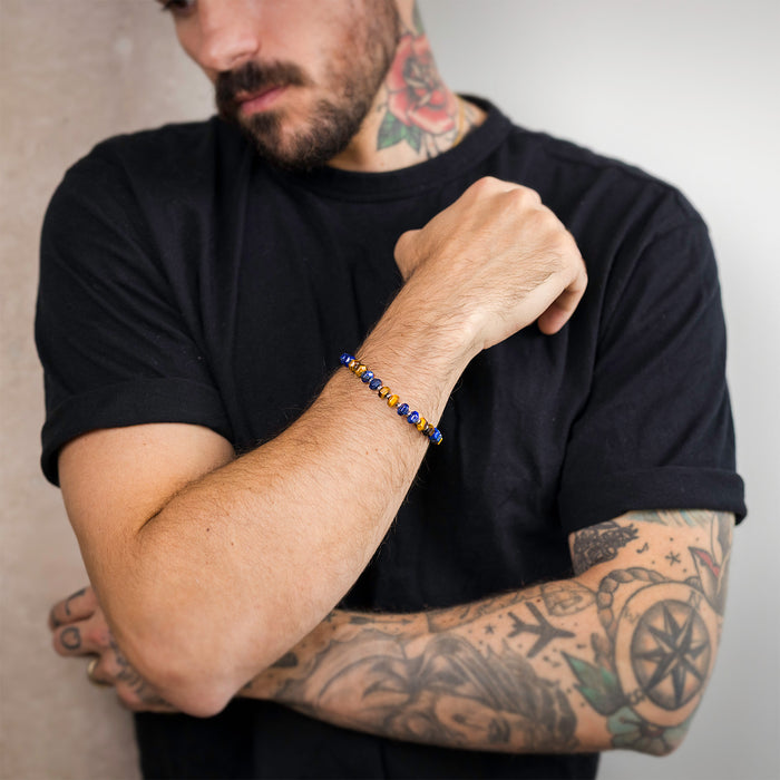 Man with tattooed arms wearing a black t-shirt and The Damascus Voyage Bracelet featuring blue and gold beads, styled as bold men's jewelry by Ralph Christian, bold by design.