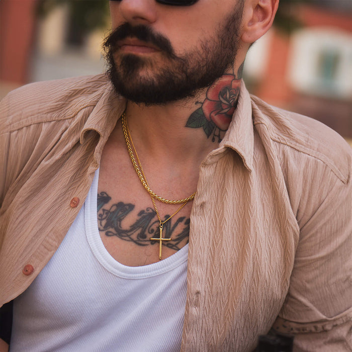 Man wearing sunglasses, a beige textured shirt over a white tank top, and The Gold Wheat Necklace - Essentials Collection, featuring a gold cross pendant on a chain, with visible neck and chest tattoos.
