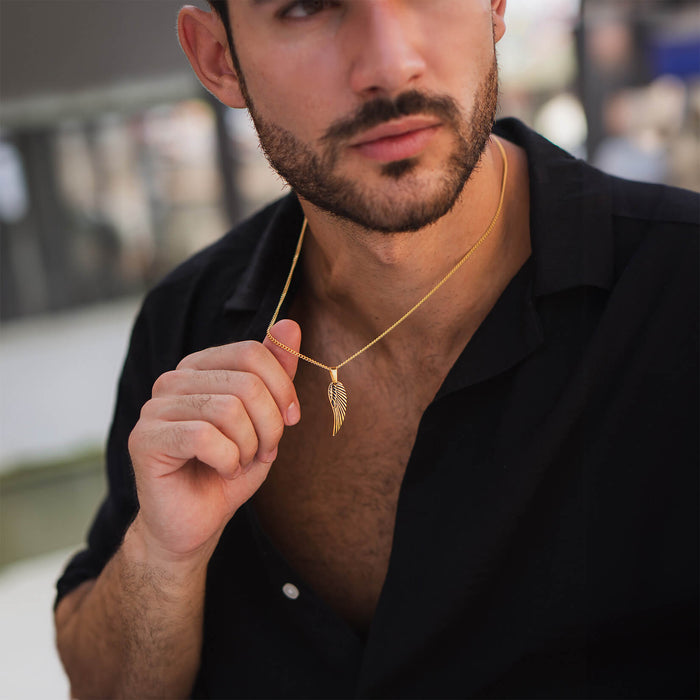 A bearded man wearing an open black shirt holding The Gold Wing Pendant - Essentials Collection, a sleek gold wing-shaped necklace designed for men.