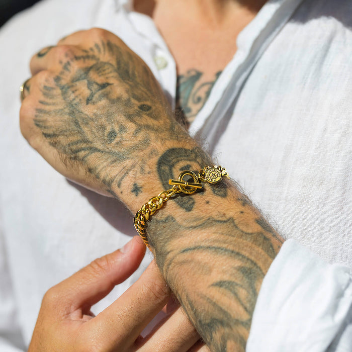 Close-up lifestyle image of a hand wearing The Medallion Bracelet - Gold, featuring a bold gold chain bracelet with a round medallion charm and toggle clasp, styled on a tattooed wrist against a white shirt, showcasing Ralph Christian's statement jewelry designed to be noticed.