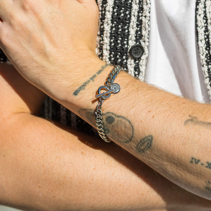 Close-up lifestyle image of a wrist wearing The Medallion Bracelet - Silver, featuring a bold silver chain design with a toggle clasp and a small engraved medallion charm, showcasing men's jewelry styled with casual attire.