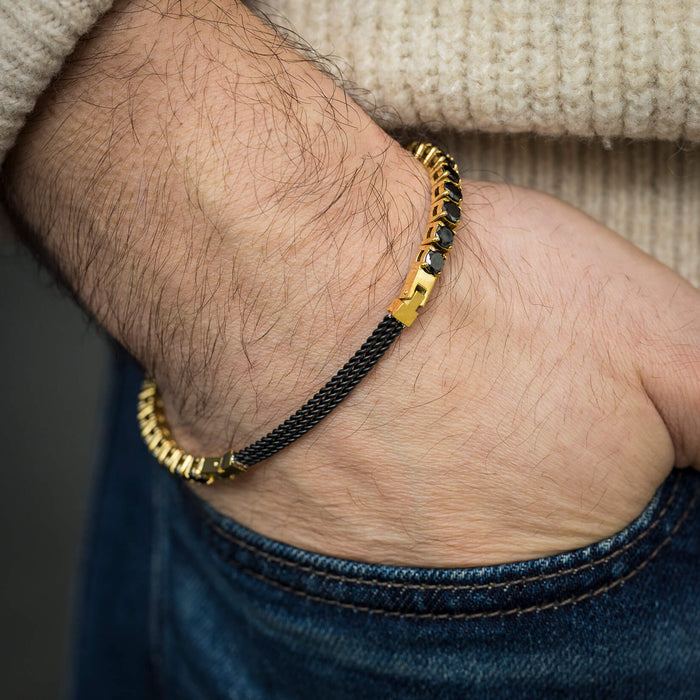 The Midnight Luxe Bracelet on a man's wrist featuring a sleek black mesh band with gold-tone accents, styled casually with a beige sweater and blue jeans.