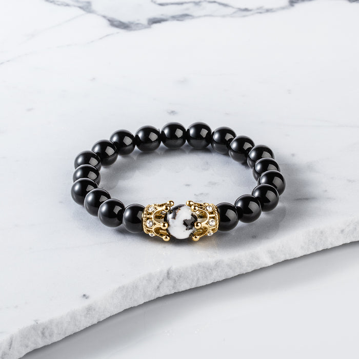 The Royal Obsidian Bracelet featuring polished black beads with a white marbled center bead framed by two gold crown accents, set on a white marble surface.