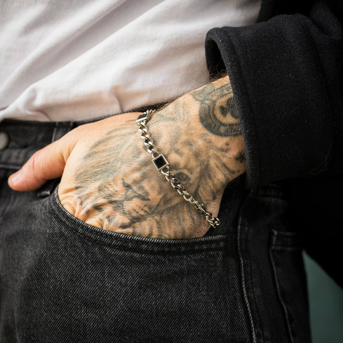 Close-up of a tattooed male wrist wearing The Urban Agate Bracelet, featuring a silver chain link design with a black agate centerpiece, styled casually with dark jeans and a black jacket.
